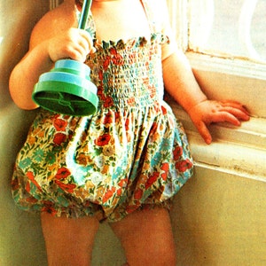 Puede incluir: Una niña pequeña con un vestido de estampado floral y un gorro a juego sostiene un juguete verde y azul. La niña está de pie cerca de una ventana.