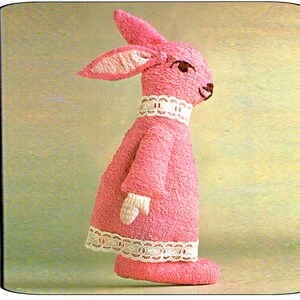 以下が含まれることがあります： 首とドレスの裾に白いレースのトリムが付いたピンクのニットのウサギのぬいぐるみ。ウサギはピンクの鼻と黒い目を持っている。