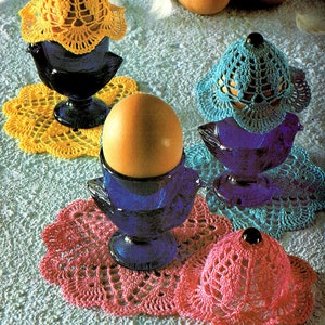 Può includere: Tre porta uova in ceramica blu con sottobicchieri all'uncinetto e coperture per uova all'uncinetto in giallo, blu e rosa.