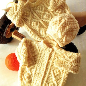 May include: A set of two knitted baby garments in a cream color. The set includes a cardigan with buttons and a matching hat. Both garments feature a textured cable knit pattern.