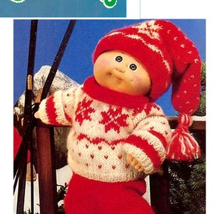 May include: A Cabbage Patch Kids doll in a red and white knitted winter outfit, including a hat, scarf, and sweater with snowflake designs. The doll is holding skis and is standing in front of a wooden fence.