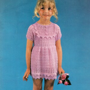 Peut inclure: Une robe en crochet violet clair avec des manches courtes et un col et un ourlet festonnés. La robe est portée par une jeune personne aux cheveux blonds et des chaussettes hautes blanches. La personne tient un petit bouquet de roses roses.