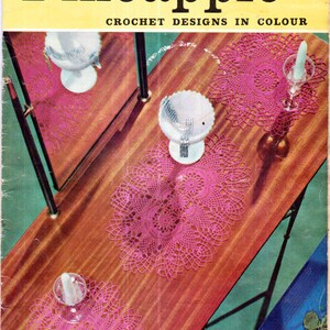 Op de afbeelding: Een vintage haakpatroonboekomslag met de titel "Pineapple Crochet Designs in Colour". De omslag toont een close-up van een houten tafel met drie roze gehaakte placemats, twee heldere glazen kaarsenhouders en een witte glazen kom met zilveren accenten.