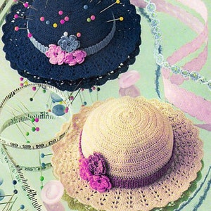 May include: Two handmade crochet pin cushions shaped like hats. One is navy blue with pink and blue floral accents and pins. The other is cream-colored with a purple band and flower. A measuring tape and pins are also visible.