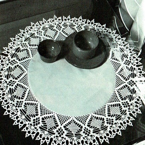 May include: White crocheted doily with a delicate lace pattern. The doily is placed on a dark surface with two bowls and a plate.