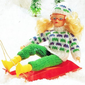 Puede incluir: Una muñeca con un suéter tejido blanco, verde y azul, pantalones tejidos verdes, un gorro tejido blanco y azul y gafas de esquí rojas. La muñeca está sentada en un trineo rojo en la nieve.