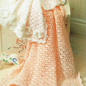 May include: Two crocheted baby blankets, one white and one peach, are folded and displayed on a white surface. The white blanket has a delicate floral pattern and a scalloped edge. The peach blanket has a lacy openwork pattern.
