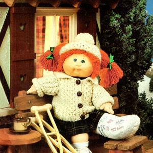 Puede incluir: Una muñeca Cabbage Patch Kid con un gorro blanco de ganchillo y un suéter de punto crema con botones marrones. La muñeca tiene el pelo de lana rojo en trenzas y está sentada en un banco de madera con muletas a su lado. La muñeca lleva una falda de cuadros y un calcetín blanco con una bota a rayas rojas y blancas. La muñeca sostiene un yeso blanco con las palabras "Get Well" y "Alison" escritas en él.