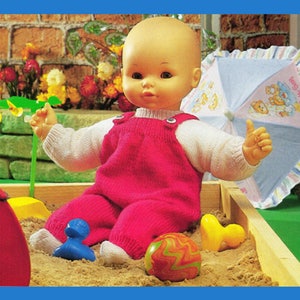 Könnte beinhalten: Eine Puppe mit einem rosafarbenen Strickoverall und einem weißen Langarmshirt spielt in einem Sandkasten. Die Puppe hat einen hellbraunen Teint und braune Augen. Die Puppe hält eine blaue Spielzeugente und einen mehrfarbigen Ball.