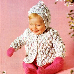 Puede incluir: Un cárdigan de punto blanco para bebé con un gorro de punto rosa y polainas rosas. El cárdigan tiene un patrón texturizado y está decorado con pequeñas flores blancas y rosas. El gorro tiene una pequeña flor en la parte superior. El bebé está sentado sobre un fondo rosa. El texto "PATONS 1267 12p Carefree D.K. & D.K.s 18-19 in. 46-48 cm" está impreso en la parte superior de la imagen.