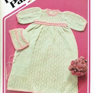 Puede incluir: Un vestido de bebé de punto blanco con ribetes rosas y un gorro de punto a juego. El vestido tiene un patrón de rombos y el gorro tiene un borde festoneado. El vestido y el gorro están colocados sobre un fondo rosa. El texto "P426 Darling 3 ply" y "Wendy design" es visible en la imagen.