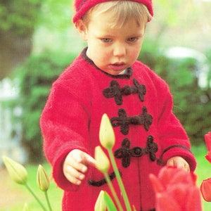 May include: A young child wearing a red knitted jacket with black buttons and a red knitted hat is standing in a garden with green foliage and red tulips.