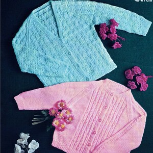 May include: Two knitted cardigans, one light blue and one pink, with button closures. The cardigans are laid out on a dark green background with flowers.