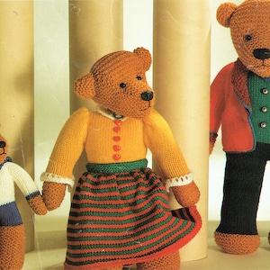 Puede incluir: Tres osos de peluche tejidos, uno con un suéter amarillo y una falda a rayas verde y roja, otro con una chaqueta roja y pantalones negros, y uno más pequeño con una camisa de marinero blanca y pantalones azules.