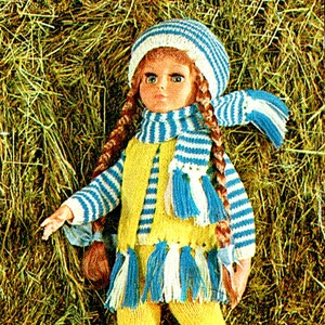 May include: A doll wearing a yellow and blue striped knitted vest, matching hat and scarf with white and blue tassels. The doll has long brown braids and blue eyes.