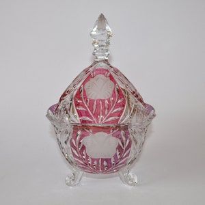May include: A clear glass candy dish with a pink and white floral design and a lid. The dish has three legs and is decorated with cut glass.