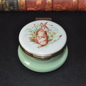 May include: A small, round, green trinket box with a hinged lid. The lid features a painted image of a brown rabbit with white markings sitting in a field of green grass and yellow flowers.