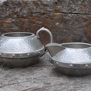 May include: A pair of antique pewter serving vessels. The set comprises a sugar bowl with two handles and a creamer with a spout. Both items feature a hammered finish and are encircled by a decorative band with rivets.