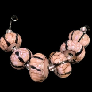 May include: A string of six round glass beads arranged in a curved line. Each bead features a pale pink base color with a textured, crackled surface pattern in black and dark brown. Small, clear spacer beads are positioned between the larger beads.