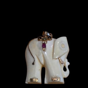 May include: An off-white elephant pendant featuring a gold-toned bail at the top. The elephant is adorned with a small cluster of multi-colored gemstones on its back, including a prominent red stone, and gold-toned accents on its feet and trunk.