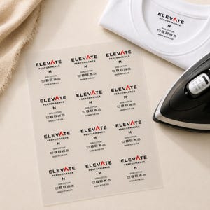 May include: A sheet of twelve iron-on clothing labels featuring the black text Elevate Performance, size M, 100% cotton, and care instructions. A white t-shirt with a matching label and a black iron are positioned nearby on a light surface.