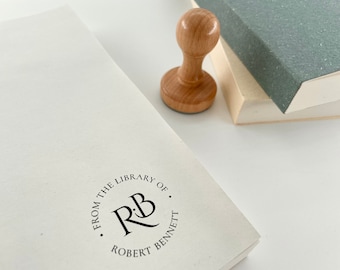 May include: A round rubber stamp with the text "From the Library of Robert Bennett" and the initials "R&B" in a circle. The stamp is on a white surface next to a wooden stamp handle.