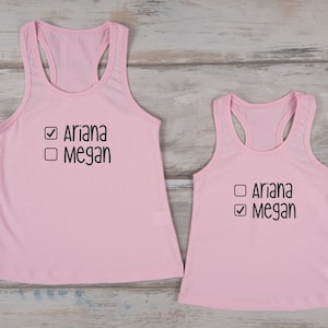 Peut inclure: Deux débardeurs roses clairs. Le plus grand débardeur est à côté d'un plus petit. Les deux ont le texte "Ariana" et "Megan" imprimé en noir, avec une coche à côté de "Megan".