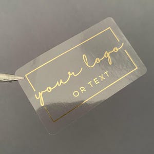 Può includere: Un adesivo rettangolare trasparente con scritte dorate e un bordo dorato. Il testo recita "your logo OR TEXT" in stile scritto a mano. L'adesivo è tenuto da una pinzetta su uno sfondo grigio.