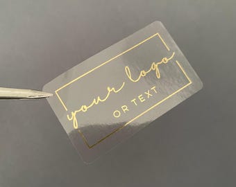Può includere: Un adesivo rettangolare trasparente con scritte dorate e un bordo dorato. Il testo recita "your logo OR TEXT" in stile scritto a mano. L'adesivo è tenuto da una pinzetta su uno sfondo grigio.