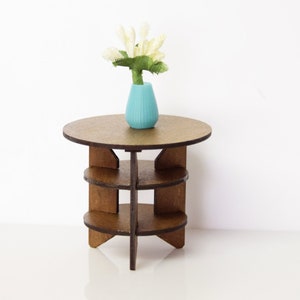 May include: A small wooden end table with three shelves. The table has a round top and a vase with white flowers on it. The vase is a light blue color.