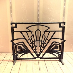 May include: Black metal fireplace screen with an Art Deco design. The screen has a geometric pattern with a central point and curved lines.