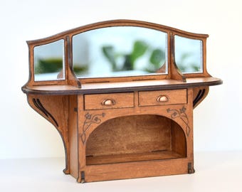 May include: A miniature wooden vanity with a central mirror and two smaller side mirrors. The piece features two drawers with small metal pulls, an open storage shelf underneath, and decorative etched floral line work on the front panels.