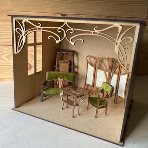 May include: A miniature dollhouse room with Art Nouveau style furniture and decor. The room features a green velvet armchair, a small table, and a screen with a floral design. The walls are painted a light brown color and the ceiling has a decorative trim.