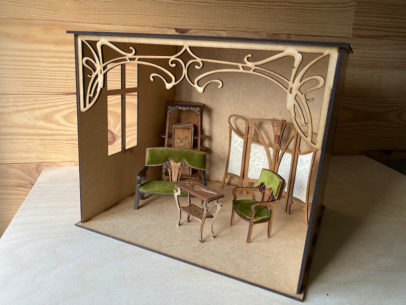 May include: A miniature dollhouse room with Art Nouveau style furniture and decor. The room features a green velvet armchair, a small table, and a screen with a floral design. The walls are painted a light brown color and the ceiling has a decorative trim.
