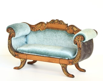 Könnte beinhalten: Ein Miniatursofa mit hellblauem Samtbezug auf Sitzfläche, Rückenlehne und den eingerollten Armlehnen. Der Rahmen besteht aus dunkelbraunem Holz und ist mit detaillierten, goldfarben bemalten Schnitzereien an den Kanten und Beinen verziert.