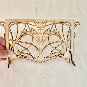 May include: A laser-cut wooden decorative panel with an Art Nouveau design. The panel features intricate swirling lines and organic shapes, with a symmetrical tree-like central motif. The panel is light brown and has a rectangular shape with curved edges.