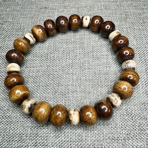 May include: A beaded bracelet featuring a mix of brown and off-white oval-shaped beads. The beads have a glossy finish and are strung together to form a circular shape. The bracelet is displayed on a gray surface.