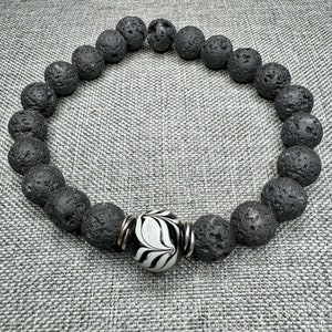 May include: A black lava rock bracelet with a white and black patterned bead. The bracelet is made of natural lava rock beads and has a silver metal clasp.