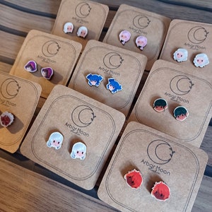 May include: A set of 10 pairs of colourful cartoon character earrings on brown card stock with the text "Arbor Moon Studios" printed on each card.