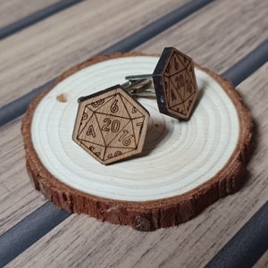 May include: A pair of wooden cufflinks, each shaped like a twenty-sided die, with etched numbers and details. The cufflinks are light brown and rest on a small, round wooden slice. The background is a wooden surface.