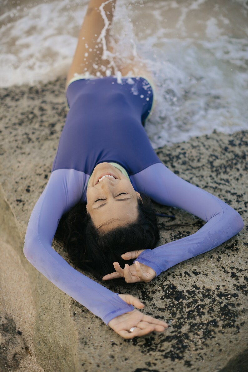 May include: A woman wearing a purple long-sleeved swimsuit with a green trim lies on a rocky beach with her eyes closed and smiling. The water is splashing around her.