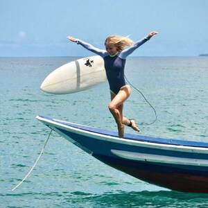 May include: A woman in a blue wetsuit jumps off a blue and white boat with a surfboard in her hand. The water is a turquoise blue.