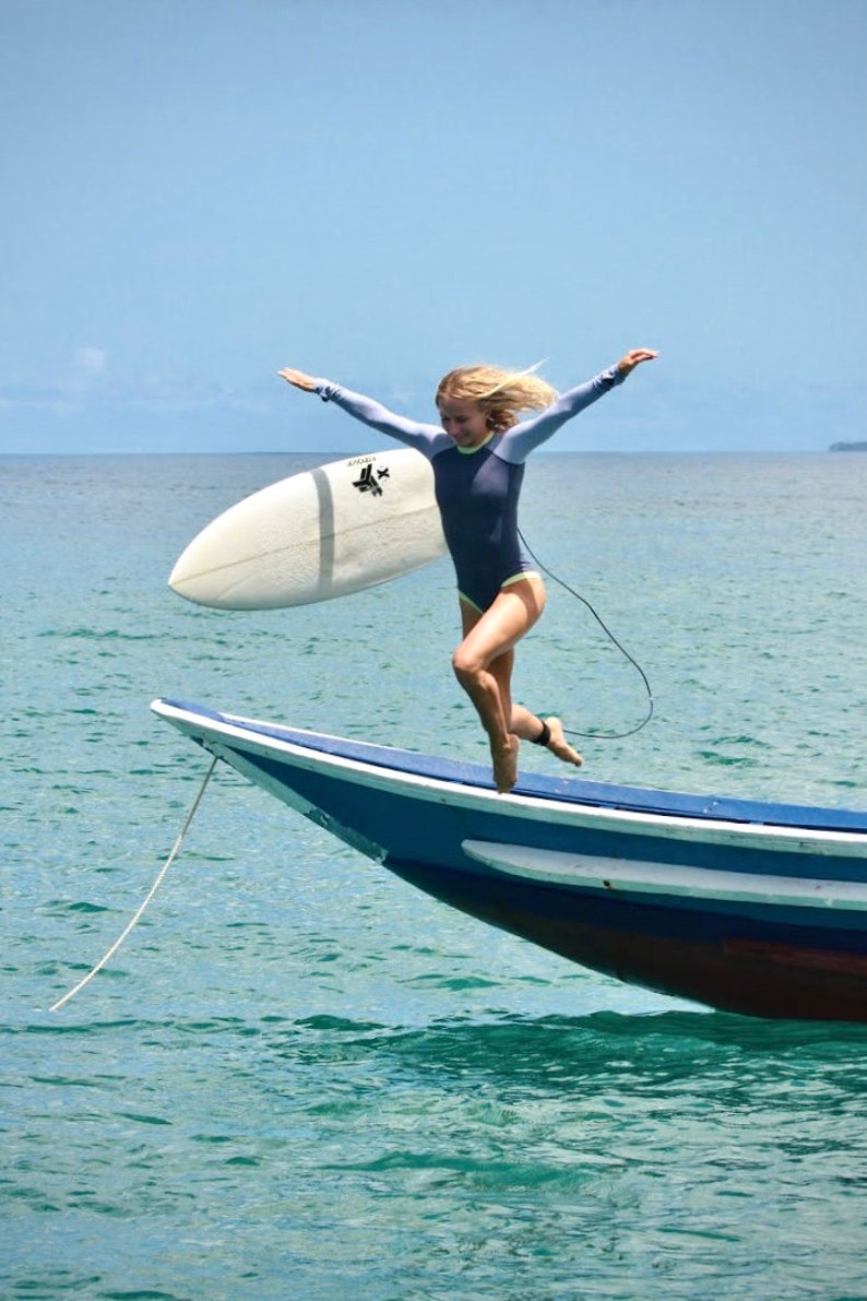 May include: A woman in a blue wetsuit jumps off a blue and white boat with a surfboard in her hand. The water is a turquoise blue.