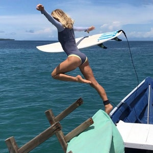 May include: A woman in a blue wetsuit jumps off a boat into the ocean with a surfboard. The boat is blue and has a wooden ladder. The ocean is a bright blue.
