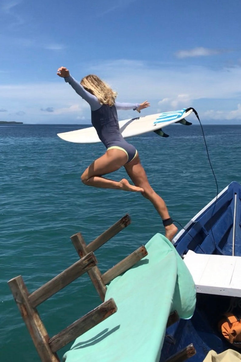 May include: A woman in a blue wetsuit jumps off a boat into the ocean with a surfboard. The boat is blue and has a wooden ladder. The ocean is a bright blue.