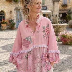May include: A long-sleeved pink blouse featuring a ruffled collar with lace trim, a button-down front, and a patchwork-style lower section. The garment includes floral patterns, polka dots, and plaid prints in various shades of pink and white.