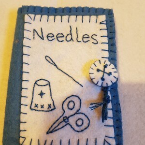 May include: A blue felt needle case with white embroidery. The embroidery includes the word "Needles", a thimble, scissors, and a needle and thread. There is a button with a cross on it.