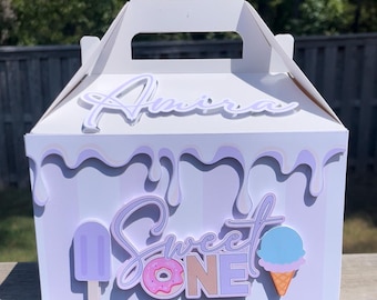 Könnte beinhalten: Eine lila und weiße Geschenkbox mit einem tropfenden Eiscreme-Design. Die Box sagt "Sweet One" und hat ein Donut-, Eiswaffel- und Eis am Stiel-Motiv. Der Name "Amira" steht auf der Oberseite der Box.