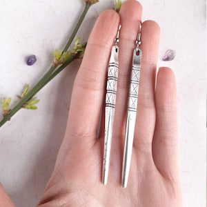 Peut inclure: Une paire de boucles d'oreilles en argent avec un motif géométrique. Les boucles d'oreilles sont longues et fines, avec une pointe pointue. Elles sont en métal et ont une finition argentée.