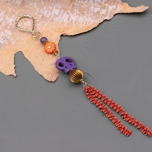 Könnte beinhalten: Ein lila Schädel-Anhänger mit orangefarbenen und goldenen Akzenten hängt an einer goldenen Kette mit roten Perlenquasten.